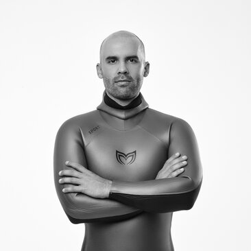 Black and white portrait of a man in a wetsuit at Amanyara, arms crossed, against neutral background.