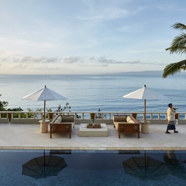Amankila Suite accommodation in Bali features a waterfront terrace with loungers, umbrellas and views across calm waters towards distant mountains, Indonesia.