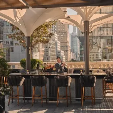 Bartender preparing cocktails at the corner bar and garden terrace at Aman New York hotel, with Manhattan skyline views.