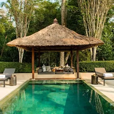 Paviliun kolam renang dengan atap jerami di Amanjiwo resort, dikelilingi taman hijau dan kursi bersantai.