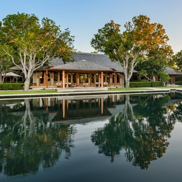 Amanyara five-bedroom villa with thatched roof reflected in still water, surrounded by tropical trees.