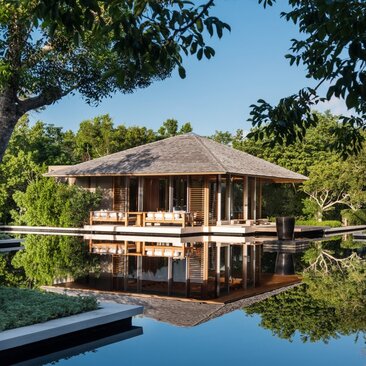 Amanyara villa's beach sala with thatched roof reflected in still pond water, framed by tropical vegetation.