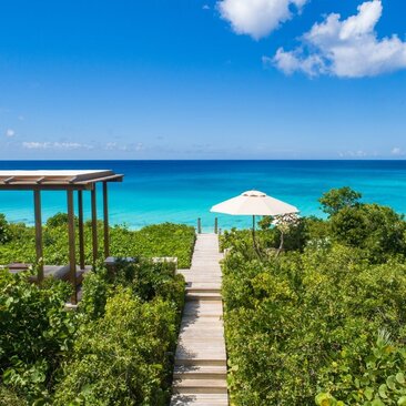 Wooden beach path lined with vegetation leading to white parasol and turquoise waters at Amanyara, Turks and Caicos.