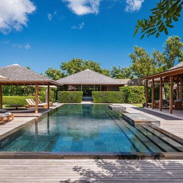 Amanyara villa with rectangular pool, wooden decking, and thatched-roof pavilions overlooking tropical gardens under clear sky.