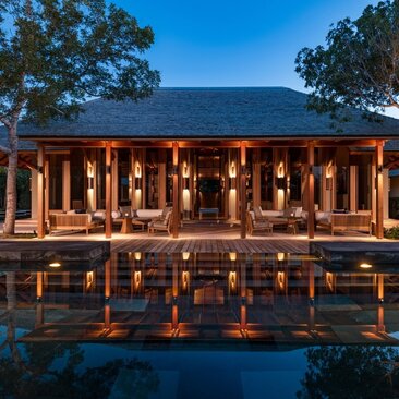 Four-bedroom villa at Amanyara illuminated at dusk, reflecting in still water with tropical trees beyond.