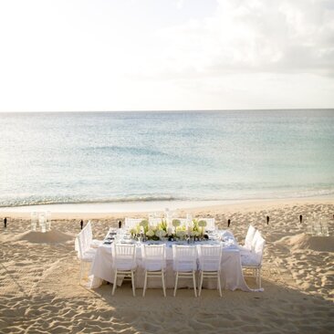 Beachside dining setup at Amanyara with a white-dressed table and chairs on sand facing turquoise waters.