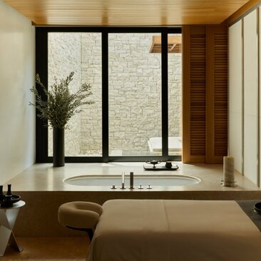 Spa treatment room at Amanzoe resort with sunken soaking tub, minimalist décor and natural light from large window.