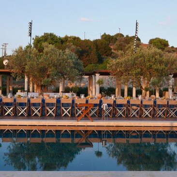 Beach club with wooden decking and classical columns overlooking still water at Amanzoe resort, Greece.