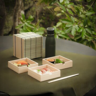 Wooden bento box with compartments of sushi and vegetables at Aman Kyoto, beside a thermos and chopsticks.