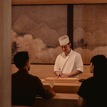 Chef demonstrating culinary techniques to two diners at Aman Nai Lert Bangkok's dining venue.