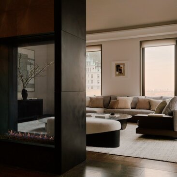 アマン ニューヨークのホテルにある、3ベッドルームの広々としたリビングエリア。大きな窓から自然光が入り、モダンなソファと暖炉が配置されています。