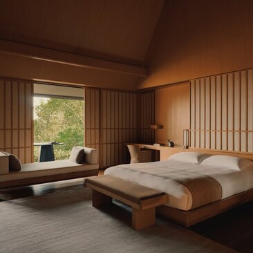 Serene principal bedroom at Amanemu resort with wooden bed, low seating area, and floor-to-ceiling windows overlooking nature.