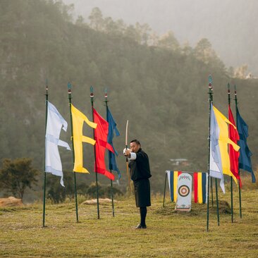 Archer in traditional dress stands beside Bhutanese and monastery flags at Amankora's Punakha Lodge archery range.