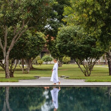 Nun in white robes sits by a reflecting pool at Amantaka, surrounded by verdant grounds and trees.
