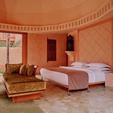Pavilion bedroom at Amanjena resort in Morocco, with terracotta walls, brass bed frame and golden velvet seating.