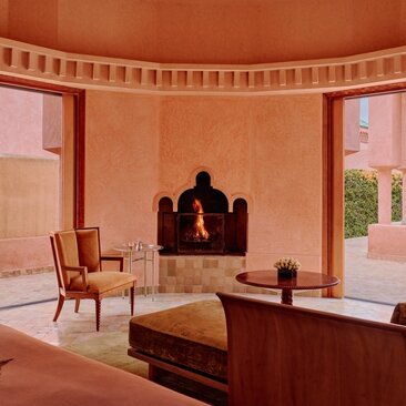 Pavilion interior at Amanjena resort with terracotta walls, wooden furniture, and fireplace opening onto Moroccan landscape.