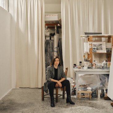 Studio portrait of Despina Flessa at Amanzoe, Greece, seated amongst art installation curtains and shelving.