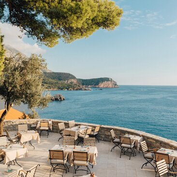 Piazza restaurant terrace at Aman Sveti Stefan, with dining chairs overlooking the Adriatic Sea and Montenegro's coastline.
