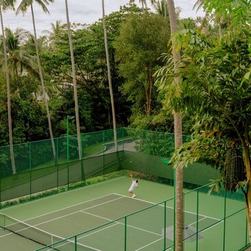 Woman playing tennis on court surrounded by lush tropical vegetation at Amanpuri, Thailand.
