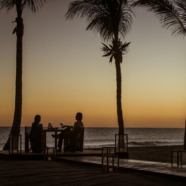 Silhouetted figures on a terrace at Amanvari, framed by palm trees against a golden sunset over water.