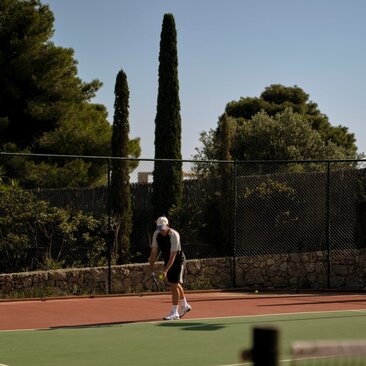 Tennis court at Amanzoe with player serving, surrounded by cypress trees and Mediterranean landscape.
