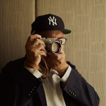 Photographer Mark de Paola holding a camera at Aman New York hotel, wearing a Yankees cap.