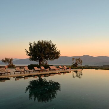 Amanzoe's main pool at sunset, with a solitary tree reflected in still water and mountains beyond.