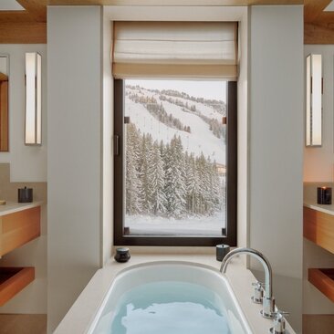 Bathroom at Aman Le Mélézin with soaking tub facing snowy alpine forest view through large window.