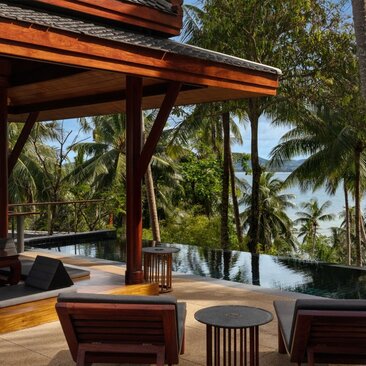 Amanpuri ocean-view pool pavilion with timber beams, teak furnishings, and tropical palms beyond.