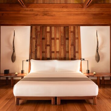 Amanpuri bedroom with wooden headboard feature, cream bedding, pendant lights, and warm timber flooring.