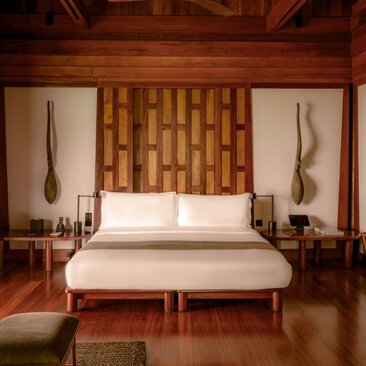 Amanpuri pavilion bedroom with teak wood headboard, cream bedding, and symmetrical bedside tables beneath carved wooden wall panels.