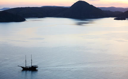 Sailing vessel anchored in calm waters at sunset, Amandira, Indonesia, with volcanic island silhouetted against twilight sky.