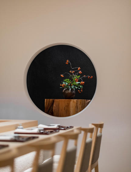 Circular black mirror reflecting a potted plant, hung above wooden dining chairs at Aman Tokyo hotel.
