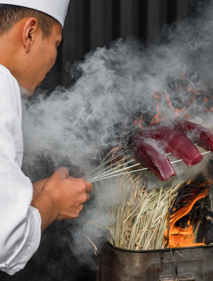 Chef grilling meat over flames at Amanemu resort, smoke rising from the grill.