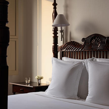 Ornate wooden bed with white linens and pillows at Amangalla, with candlelit bedside table and arched doorway.