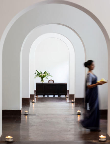 Arched corridor at Amangalla with polished grey stone floor, white walls, and a figure walking past a potted plant on a dark console table.