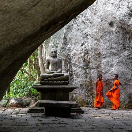 Stone steps descend beneath a cave overhang at Amangalla, with two monks in orange robes standing nearby.