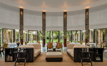 Amansara's curved reception hall with floor-to-ceiling windows overlooking gardens, featuring dark timber columns and contemporary furnishings.