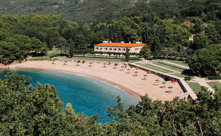 Aerial view of Aman Sveti Stefan's sandy beach cove with turquoise waters and orange-roofed buildings nestled among green hillside.