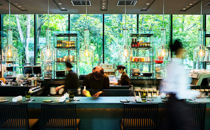 Aman Tokyo's dining café with floor-to-ceiling windows overlooking greenery, featuring a sleek counter and suspended pendant lights.
