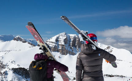 Two skiers holding their equipment at Amangani, with snow-covered peaks beyond.