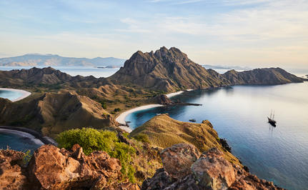 Aerial view of Amandira's rugged coastline with forested peaks meeting calm blue waters and distant mountains.