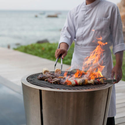 Person grilling fresh catch over an open flame at Amanoi, with coastal views beyond.