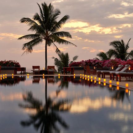 Amanpuri's waterfront at dusk, with palm trees reflected in still water and warm lights illuminating the resort buildings.