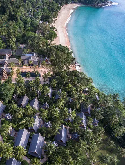 Aerial view of Amanpuri resort nestled along a turquoise coastline with sandy beach and forested grounds.