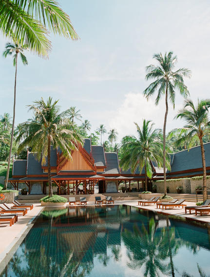 Amanpuri pool surrounded by palm trees and wooden pavilions at sunset.