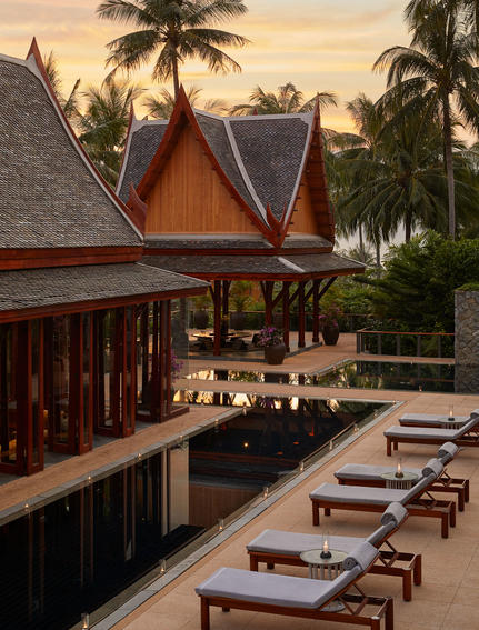 Amanpuri dining pavilion at sunset, with table settings arranged on a wooden deck beneath traditional Thai architecture and palm trees.