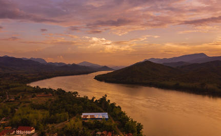 Amantaka overlooks the Mekong River at sunset, with golden light reflecting across the water and forested hills beyond.