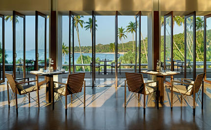 Amanwella dining venue with floor-to-ceiling windows overlooking coastal views and wooden chairs at tables.
