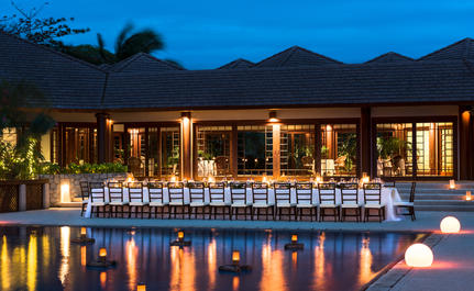 West Villa clubhouse at Amanpulo with dining area overlooking illuminated pool at dusk.